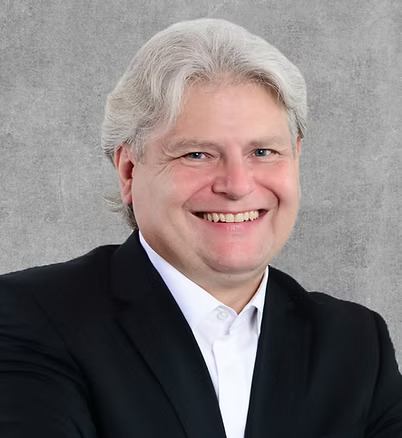 A close-up headshot of a man in a black suit and a white shirt, smiling.